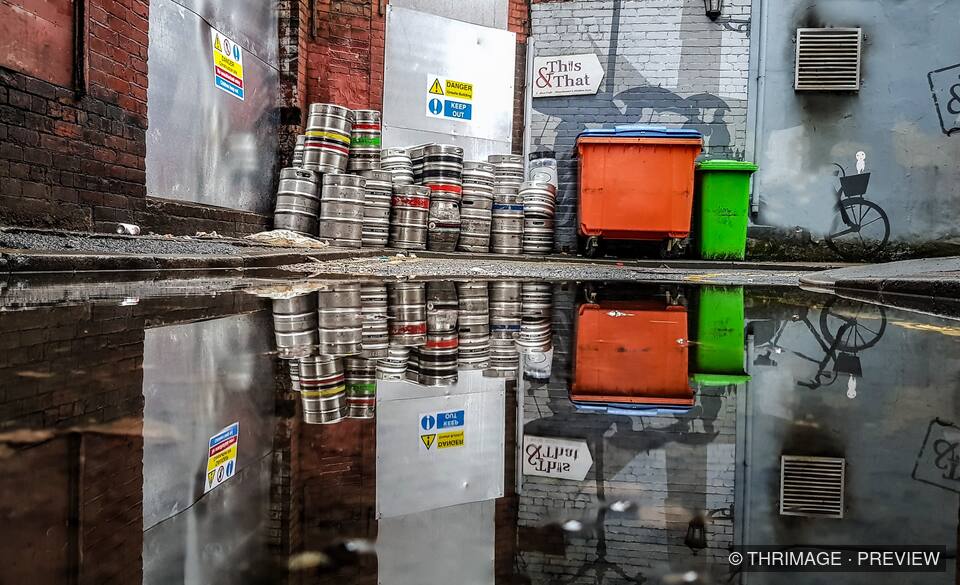 Beer kegs and orange and green wheelie bins reflected in a puddle beside brick and painted walls