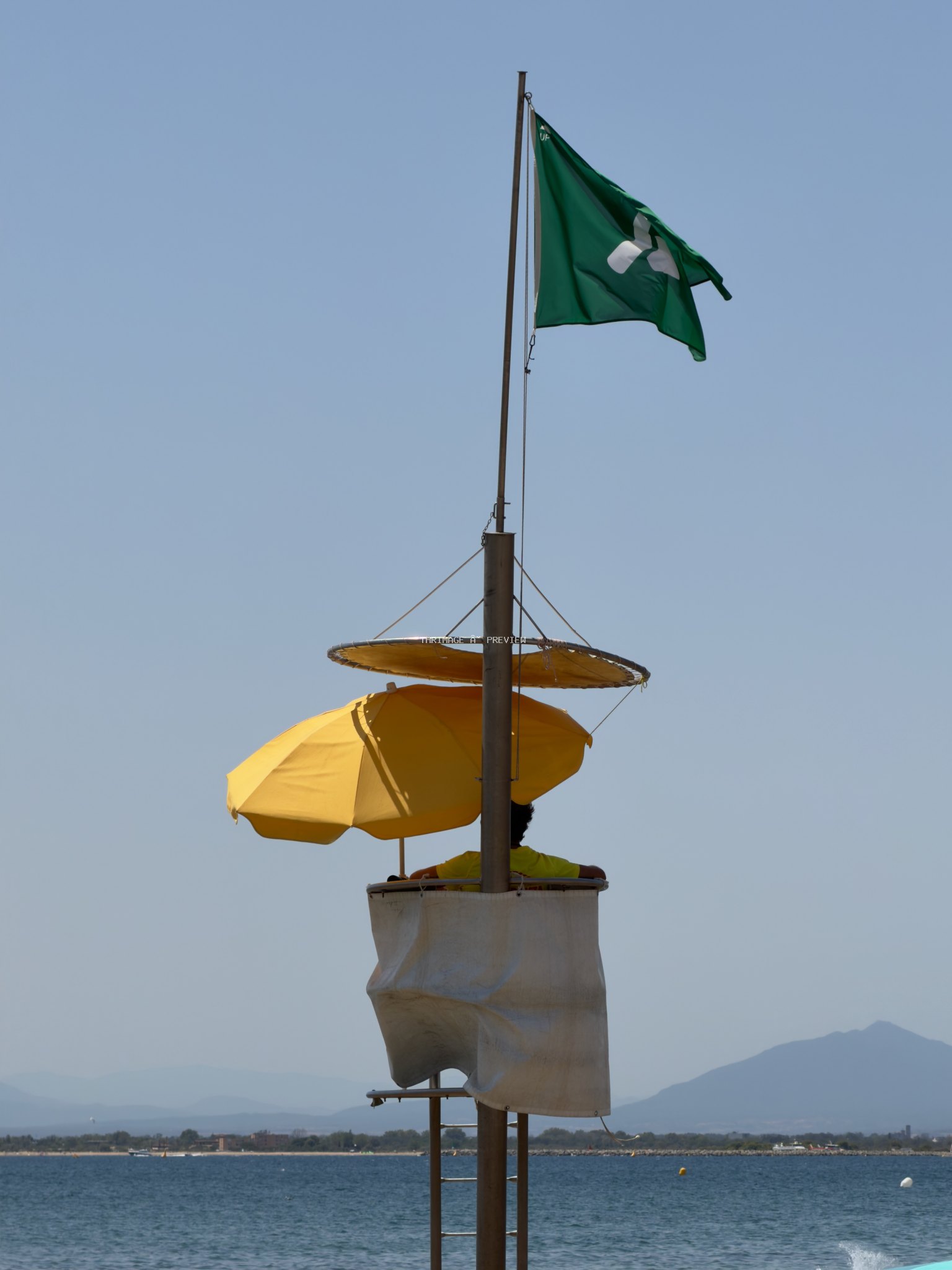 Lifeguard tower with a green flag and yellow umbrellas beside calm water, with distant mountains under a clear sky.