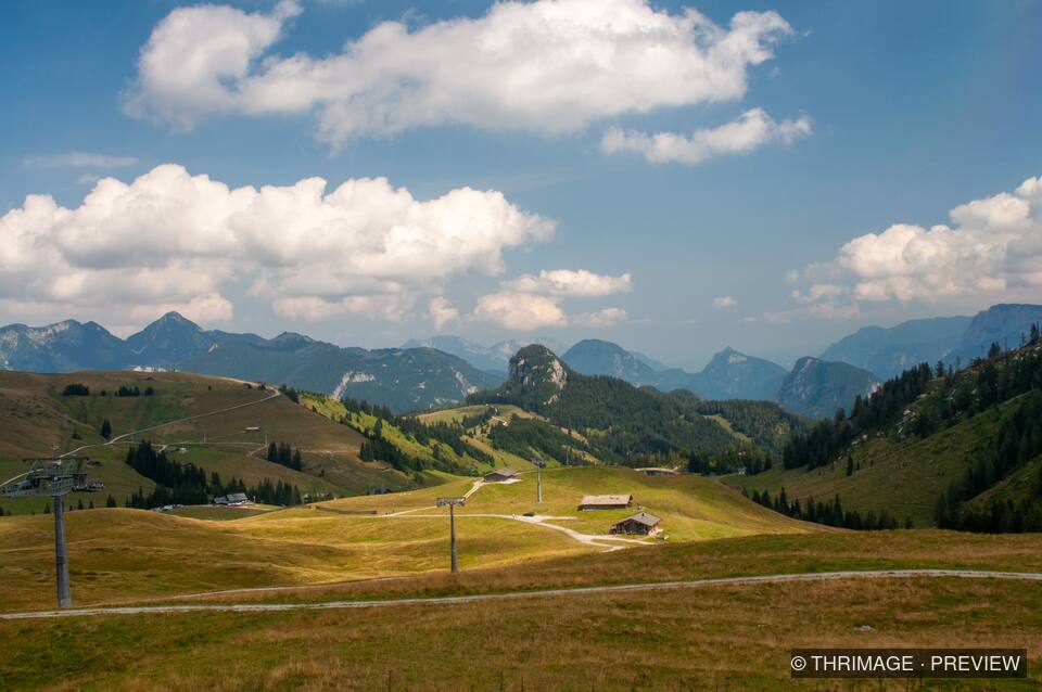 Alpine valley with grassy meadows, chairlifts, forested slopes, and rugged mountain peaks beneath white clouds.