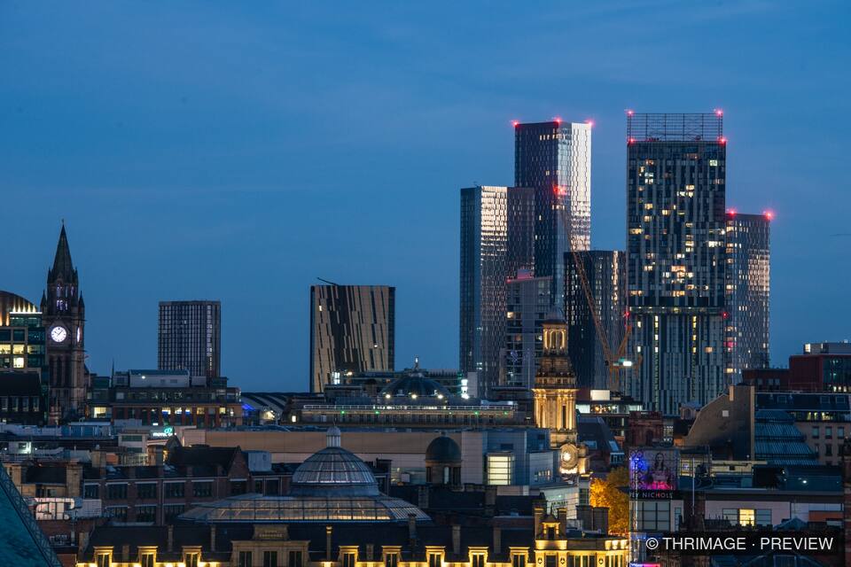 Manchester skyline at blue hour with illuminated skyscrapers, historic buildings and a prominent clock tower.