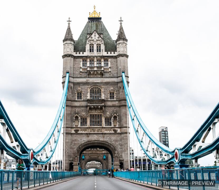 Symmetrical view through a Tower Bridge tower with blue suspension cables, roadway, and pedestrians under an overcast sky.