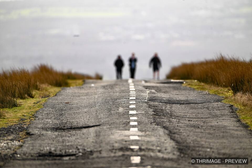 Three blurred walkers on a worn road through brown moorland beneath a hazy cloudy sky.