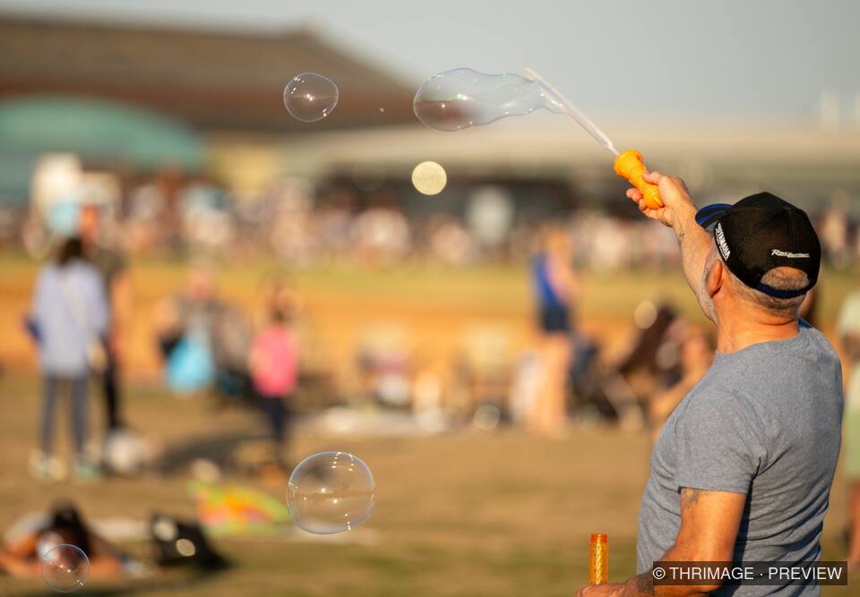 Man in a cap creating large soap bubbles with a wand at an outdoor gathering.