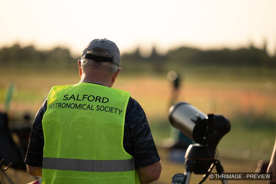 Volunteer in a Salford Astronomical Society vest stands beside a telescope in a sunlit field.