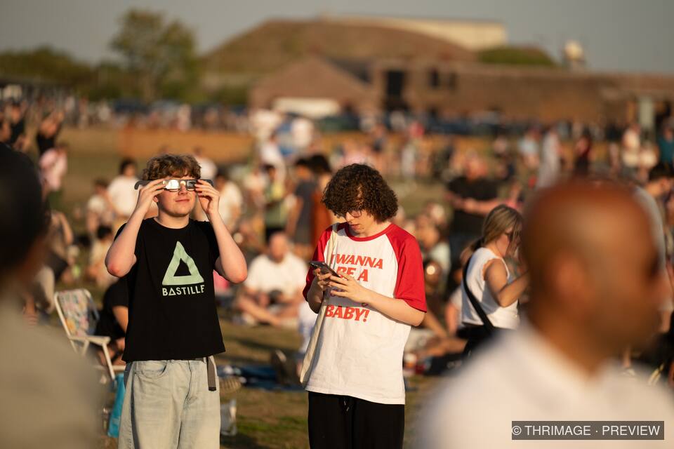 Young visitor wearing eclipse glasses looks upward while another checks a phone in a crowded outdoor gathering.