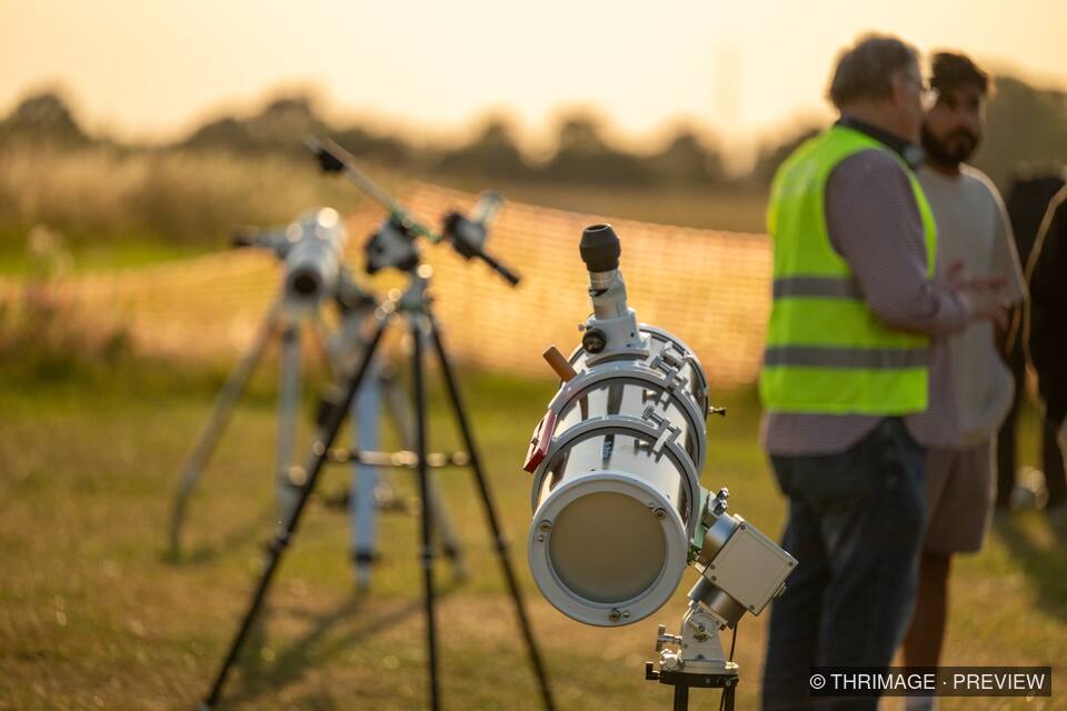 Telescope on a mount in the foreground with other telescopes and people gathered outdoors at sunset.