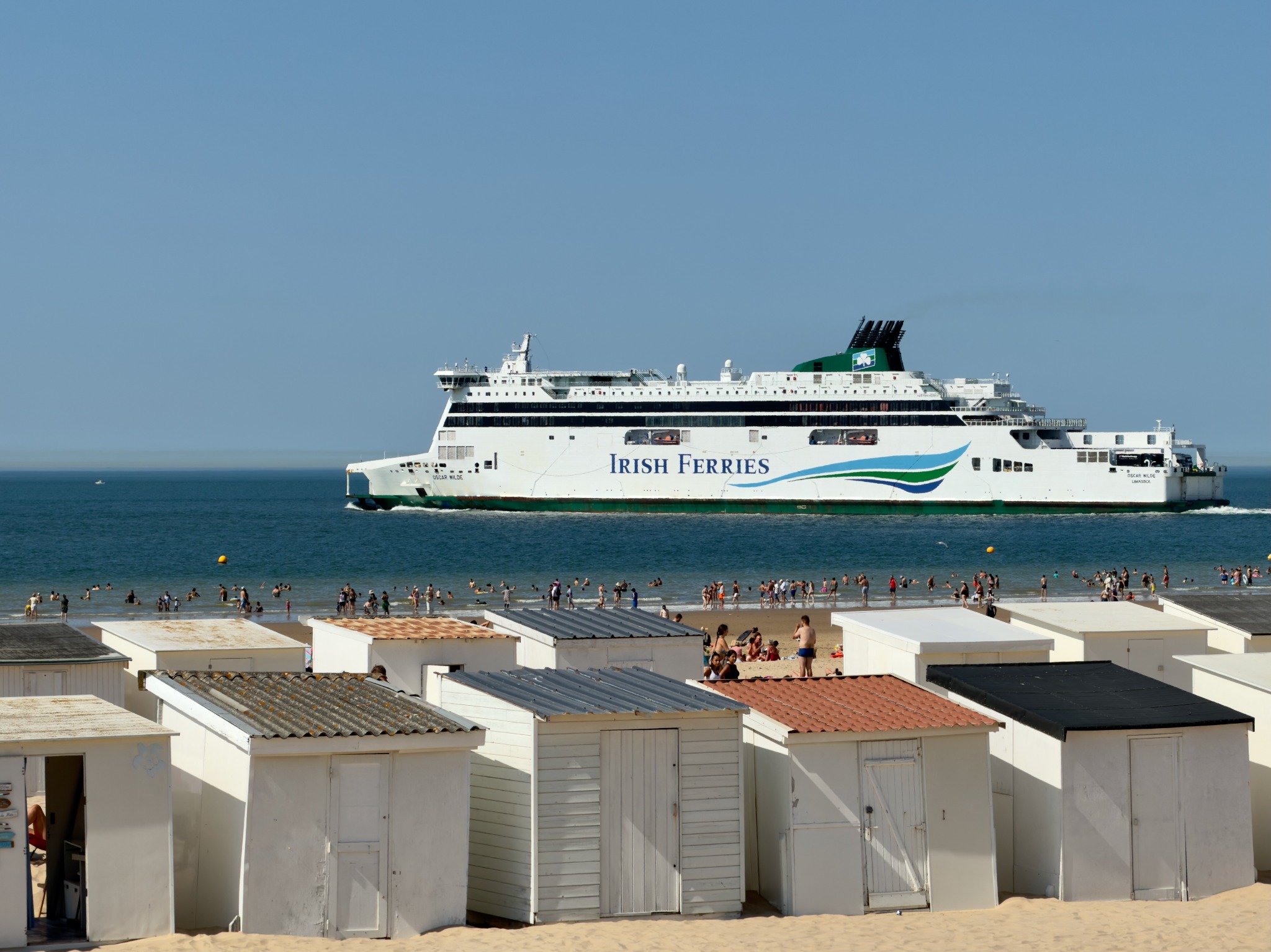 Large Irish Ferries passenger ship passing swimmers and white beach huts on a sunny beach.