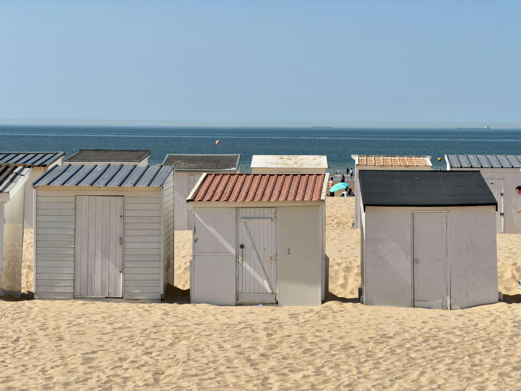 White beach huts on sand overlooking a calm blue sea with distant ships