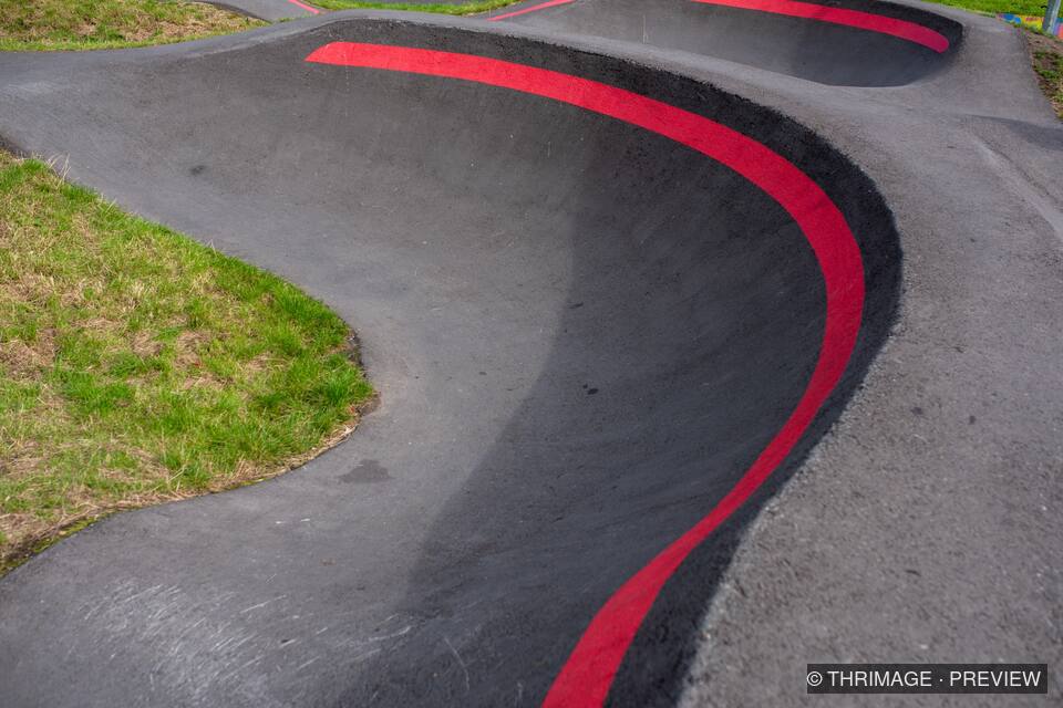 Curving black asphalt pump track with a bold red line and grassy edges